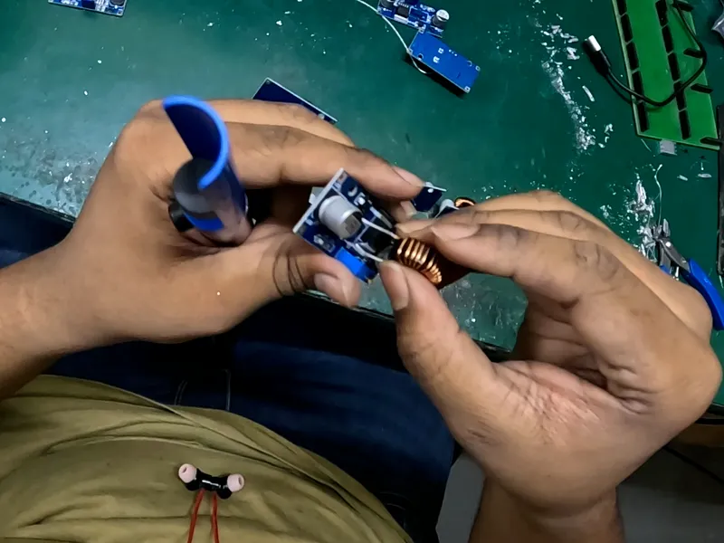Head-camera view of two hands fitting a copper coil onto a small circuit board, a desoldering pump held in the left hand.